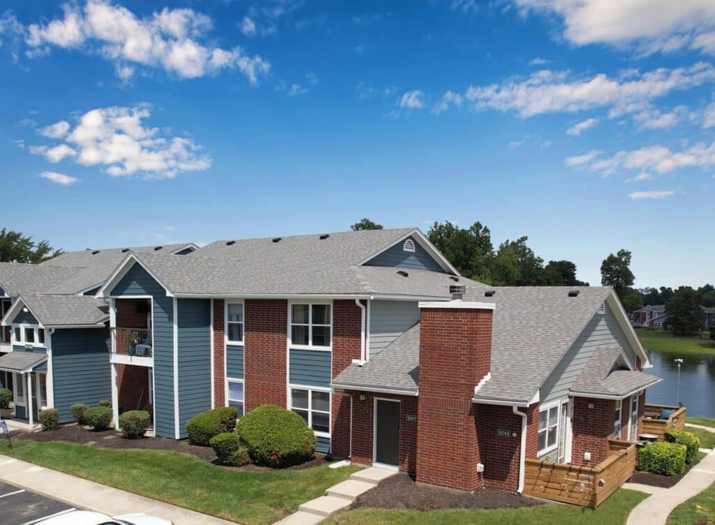 A two-story apartment building with red brick and blue-gray siding, surrounded by shrubs, under a blue sky with scattered clouds, and a pond visible in the background.