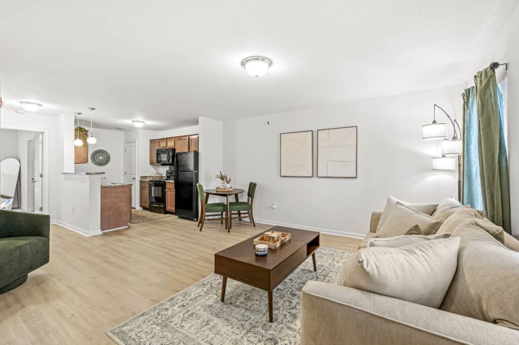 A modern open-concept living and dining area with light wood flooring, neutral furnishings, and a kitchen with black appliances in the background.