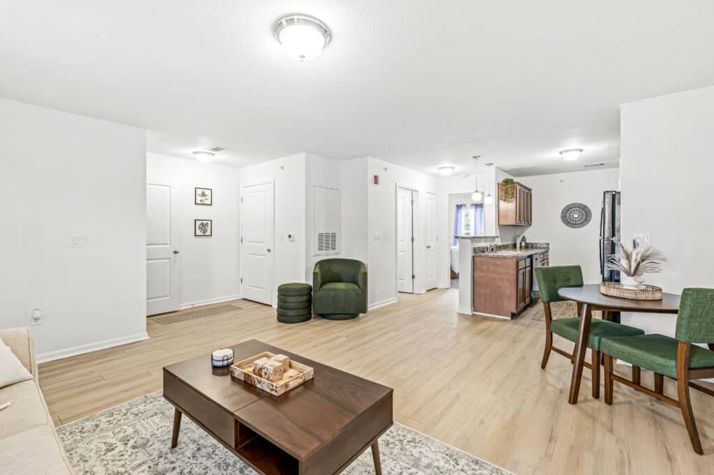 A modern apartment interior with light wood flooring, white walls, a dining table with green chairs, a coffee table, and a view into the kitchen area.