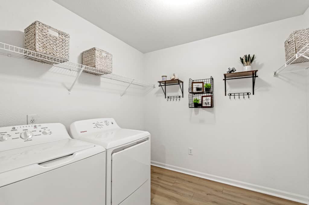 A laundry room with a white washer and dryer, wire shelves holding wicker baskets, and wall-mounted décor with small plants and accessories on a light wood floor.