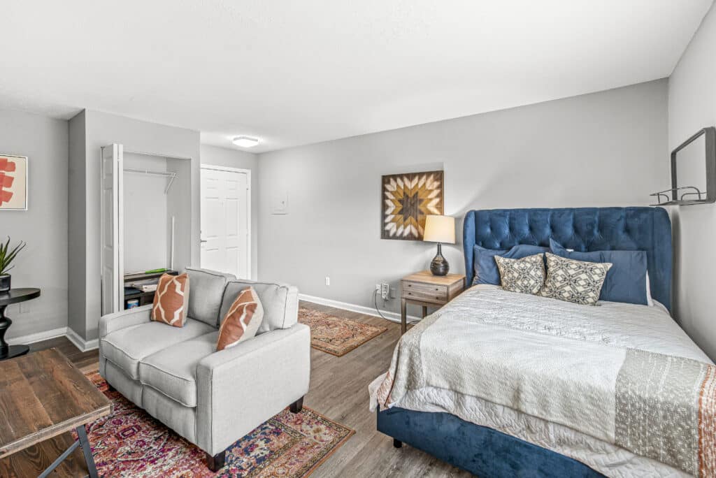A modern studio apartment with a blue upholstered bed, gray loveseat, wooden bedside table with lamp, open closet, and light-colored walls and flooring.