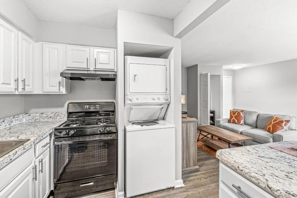 Compact kitchen with a black stove, white cabinets, marble countertops, and a stacked washer and dryer unit adjacent to an open living room with a gray sofa and orange pillows.