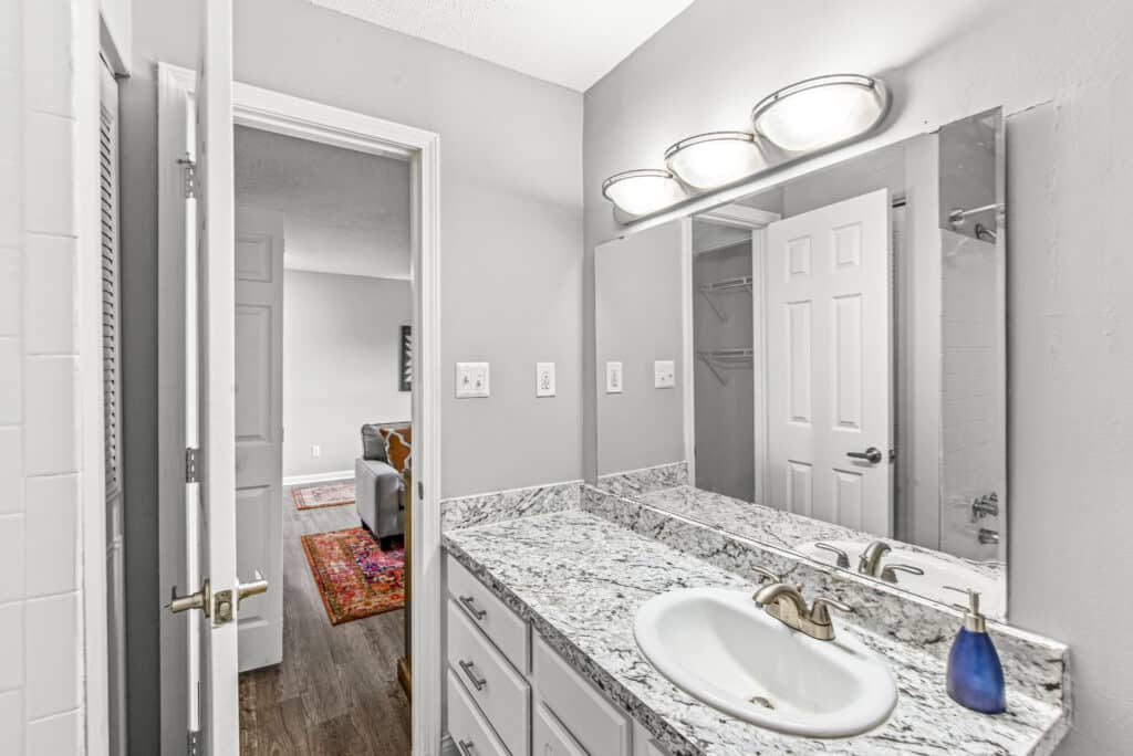 Modern bathroom with granite countertop, white cabinets, sink, wall-mounted mirror, overhead lights, and a blue soap dispenser; open door leads to bedroom with wood flooring and a red rug.