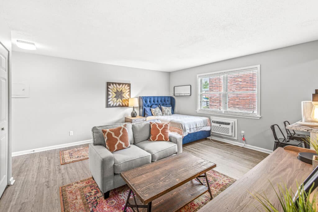 A studio apartment with a bed, sofa, wooden coffee table, desk, and window with blinds. The space features wood flooring, neutral walls, and minimal decor.