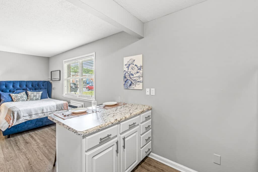 A bedroom with a blue bed, two pillows, and a blanket is adjacent to a kitchen counter with two place settings. Light gray walls and wood flooring are visible.