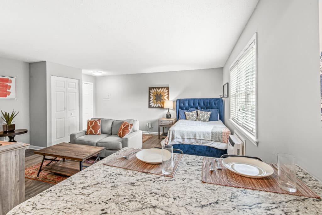 Open studio apartment with a bed, sofa, coffee table, and a kitchen counter set for two, featuring neutral walls and natural light from a large window.