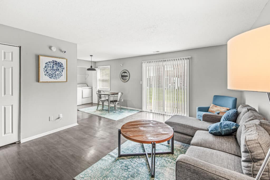 Modern living room with gray sectional sofa, round wooden coffee table, blue armchair, and open dining area; sliding glass doors lead to the outside, and laundry area is visible.
