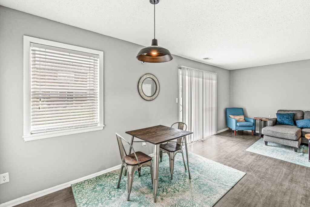 A small dining area with a wooden table and two metal chairs, a round wall mirror, and a hanging light fixture; adjacent to a living space with teal chairs.