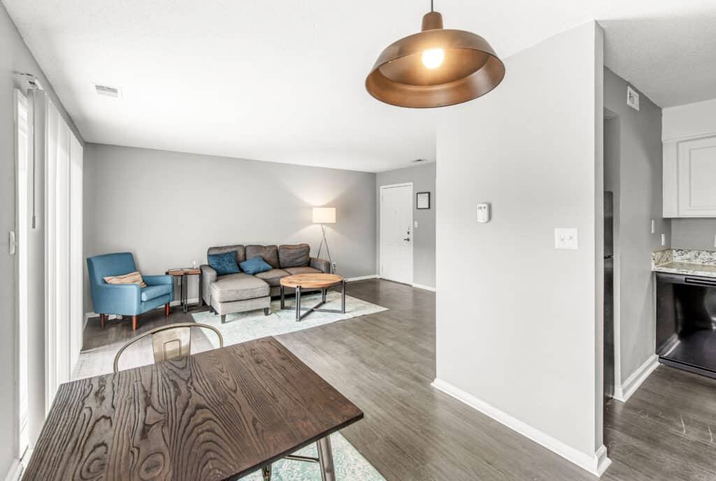 Modern living room with gray walls, wooden floors, blue and gray furniture, a round coffee table, and a dining table in the foreground under a pendant light.