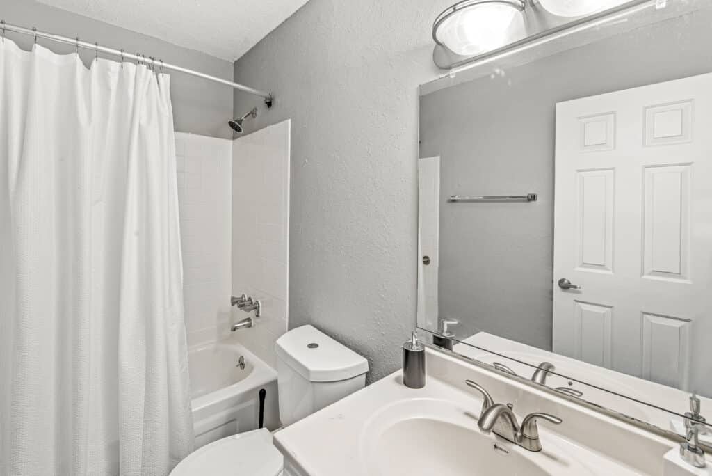 Modern bathroom with a white shower curtain, bathtub, toilet, sink with a silver faucet, and a large mirror above the vanity. Gray walls and a towel rack are also visible.
