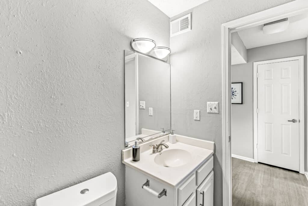 Bathroom with a white vanity and sink, wall-mounted mirror, overhead lights, soap dispenser, and a glimpse into an adjacent hallway with a closed door.