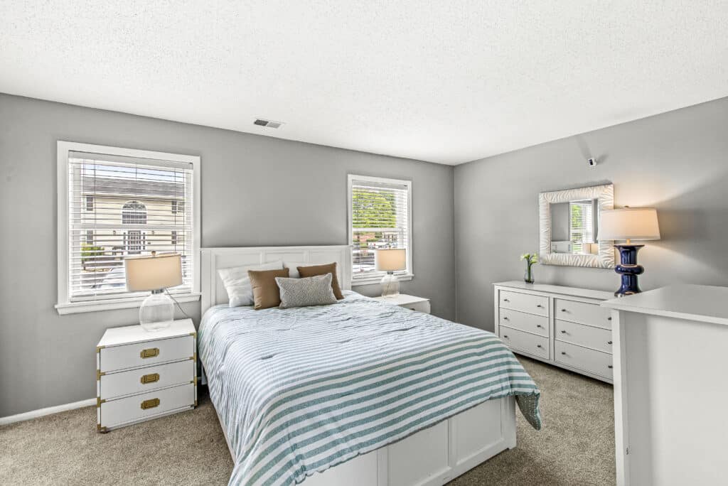 A bedroom with a bed, striped bedding, two windows, two bedside tables with lamps, a dresser with a mirror, and neutral-colored walls.