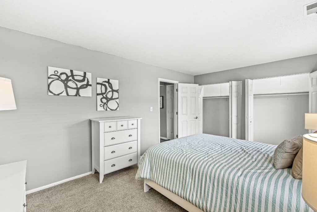 Bedroom with a striped bedspread, white furniture, two abstract art pieces on the wall, open closet doors, and neutral-colored walls and carpet.