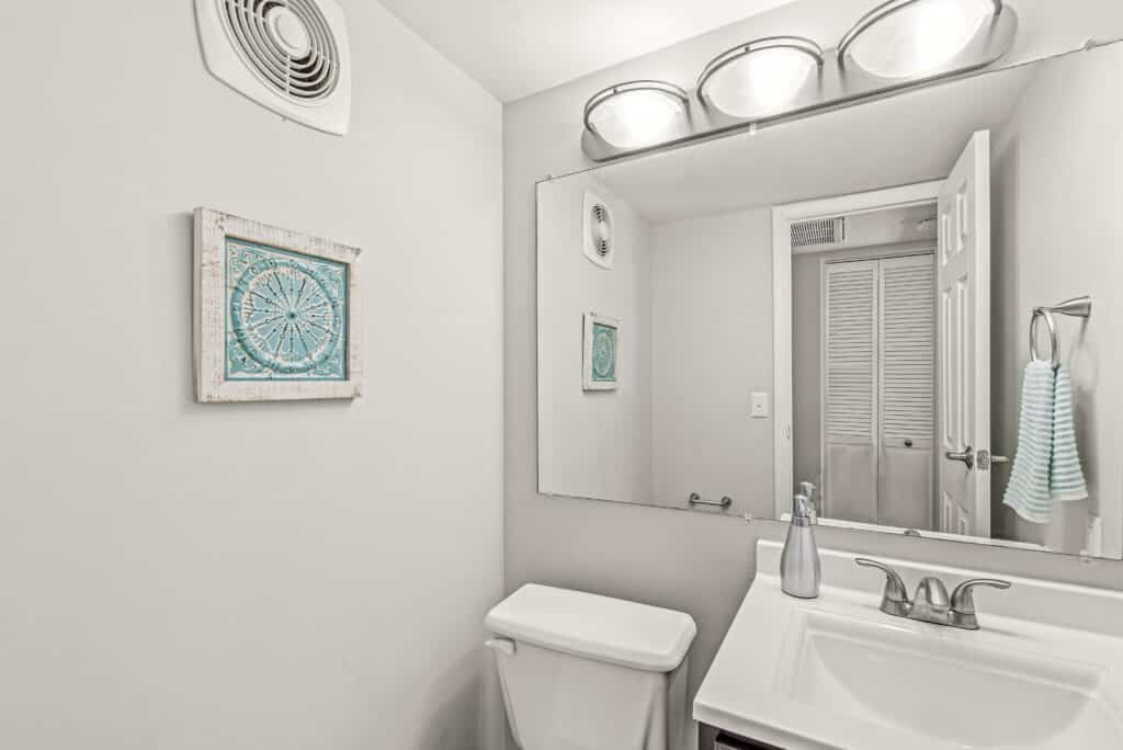 A small bathroom with a white sink, silver faucet, mirror, toilet, wall art, towel ring with striped towel, and overhead lighting.