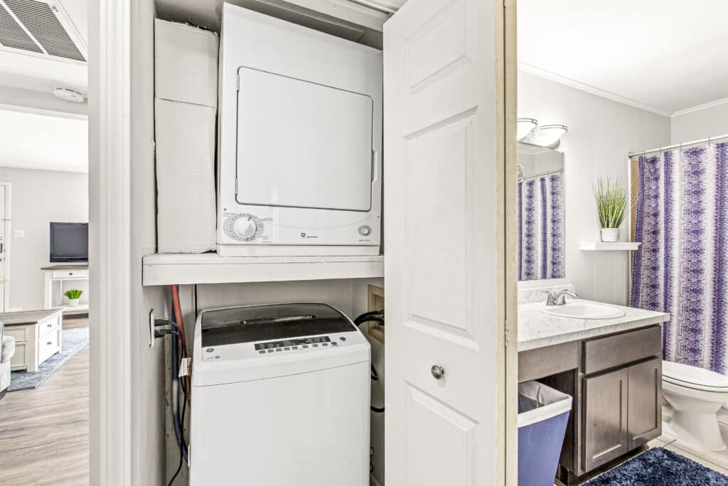 Stacked washer and dryer unit in a closet adjacent to a bathroom with a vanity, purple shower curtain, and decorative plant.
