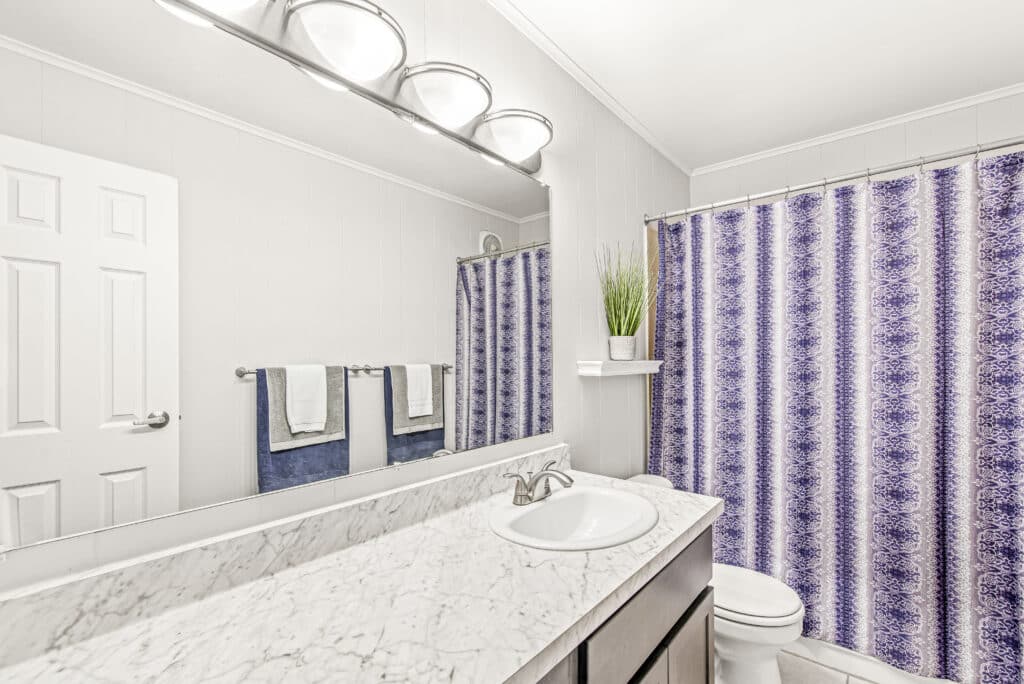 Bright bathroom with a marble countertop, single sink, large mirror, purple patterned shower curtain, two towels on a rack, and a potted plant on a white shelf.