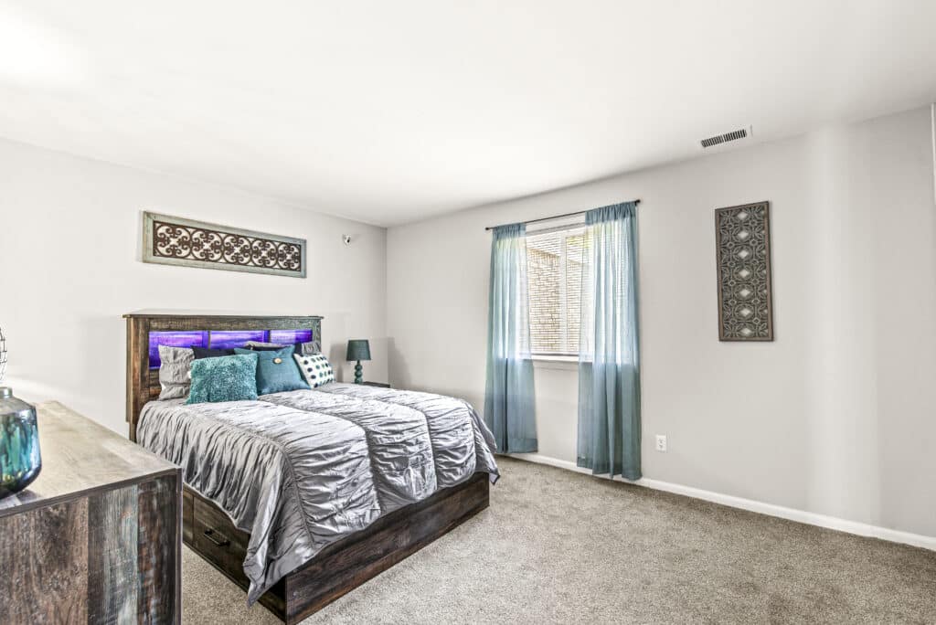 A bedroom with a bed, blue and silver bedding, matching curtains, decorative wall art, carpeted floor, and natural light from a window.