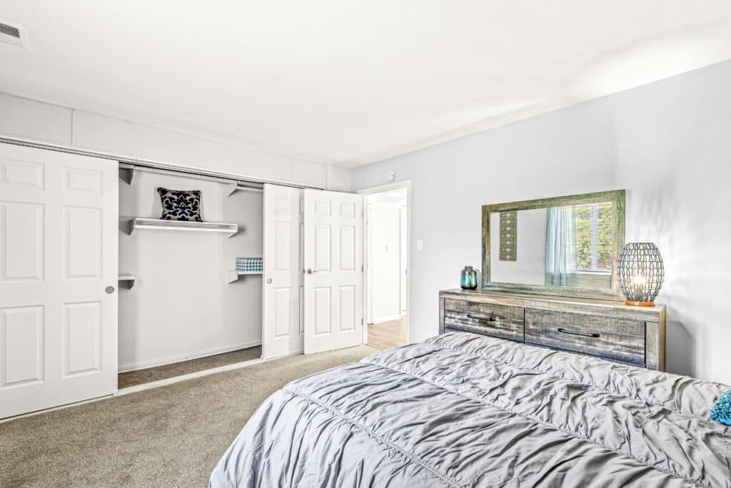 A bedroom with a bed, a dresser with a large mirror, a lamp, and an open closet with shelves and closed sliding doors. The walls are light-colored and carpet covers the floor.
