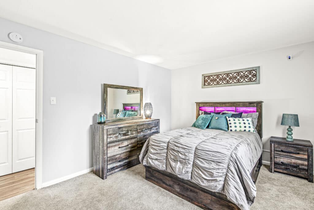 A bedroom with a wooden bed, gray bedding, nightstand, lamp, dresser with mirror, and wall art above the headboard. The room has light walls and carpeted flooring.