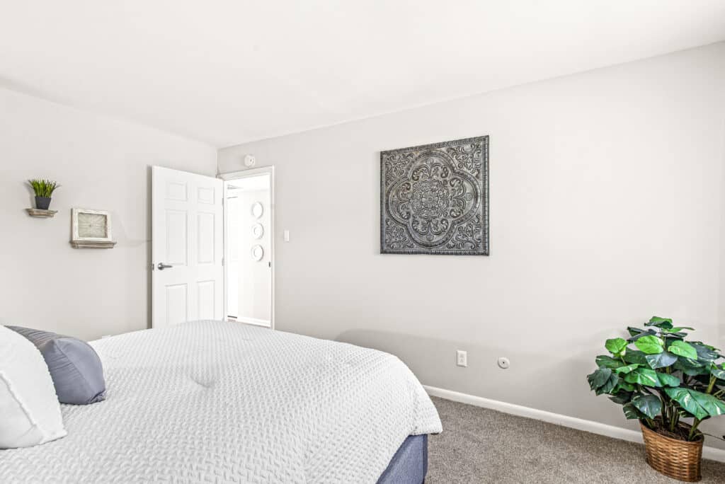 A neatly made bed in a bright bedroom with neutral walls, a large decorative wall art, a potted plant, and an open door leading to a hallway.