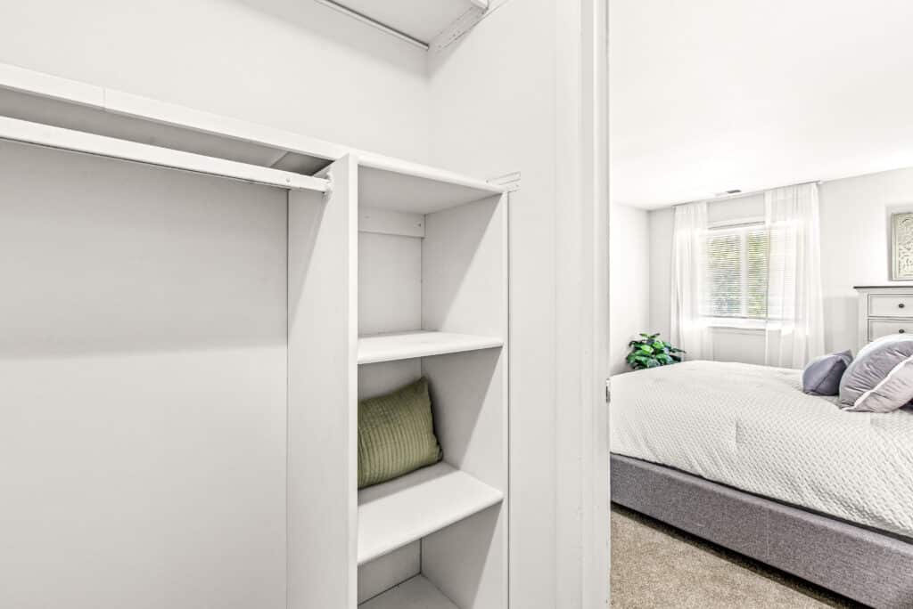 Open closet with white shelves and a green pillow in the foreground, and a bedroom with a bed, pillows, and large window visible in the background.