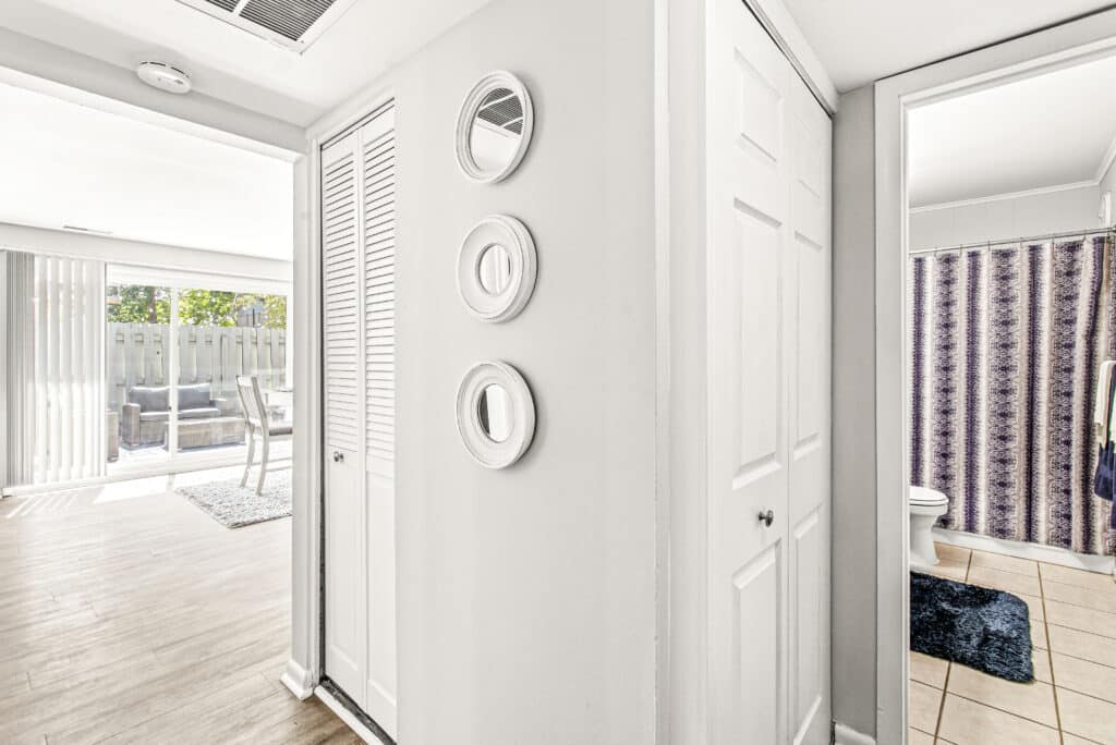 A bright hallway with white walls and three round mirrors, leading to a living area with sliding glass doors and a bathroom with a patterned shower curtain.