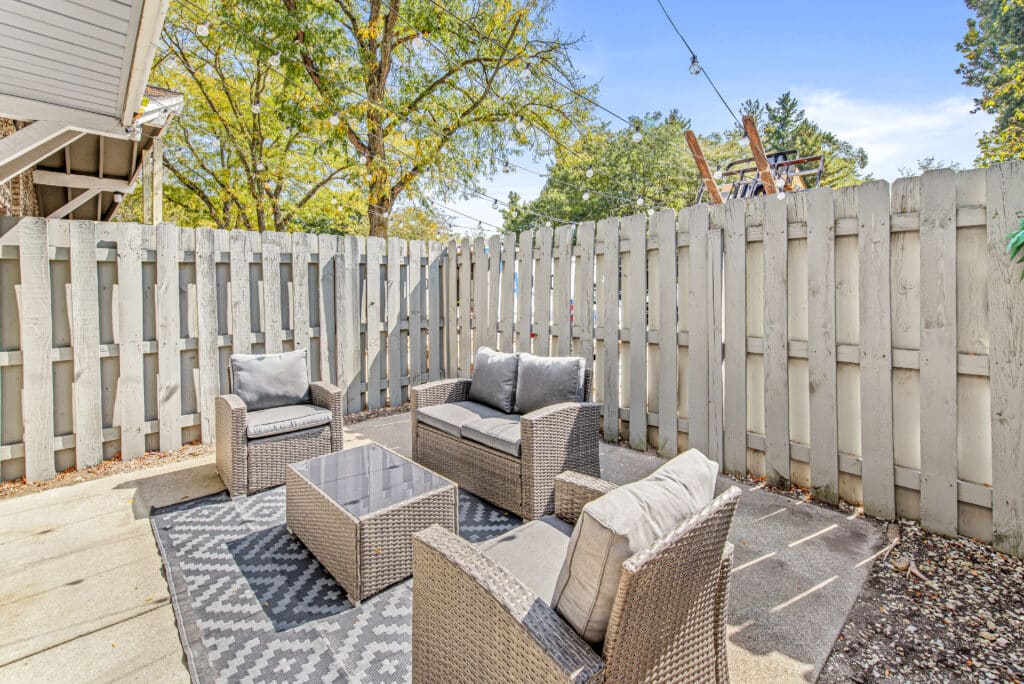 Outdoor patio area with four wicker chairs, a coffee table, and a patterned rug, enclosed by a white wooden fence and surrounded by trees under a clear sky.