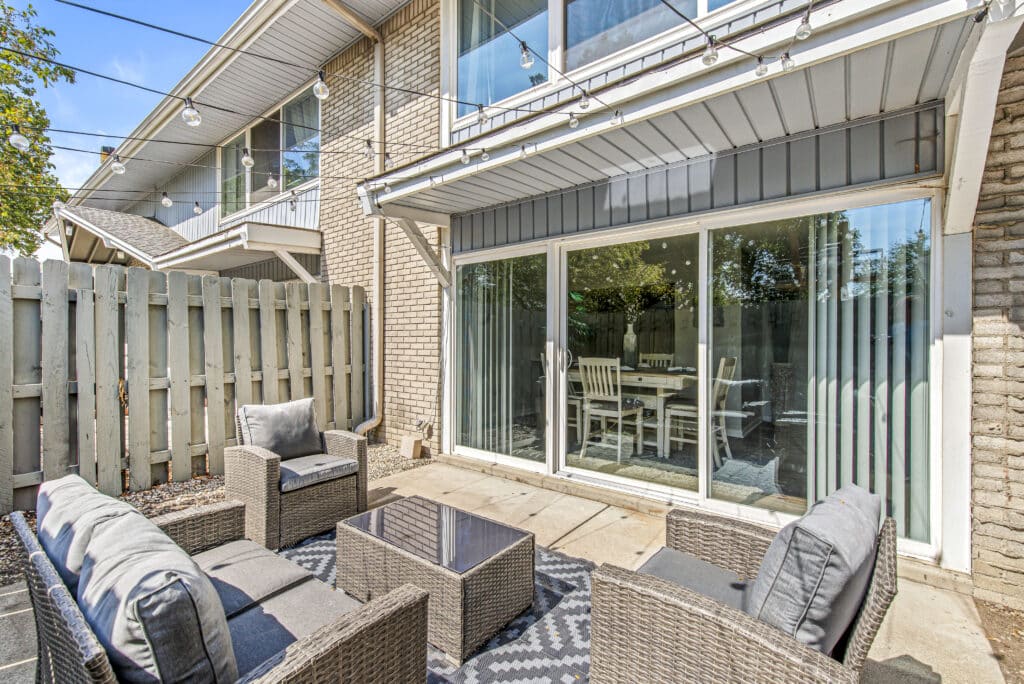 Outdoor patio area with wicker seating, a glass-top table, string lights overhead, and sliding glass doors leading to a dining room inside.