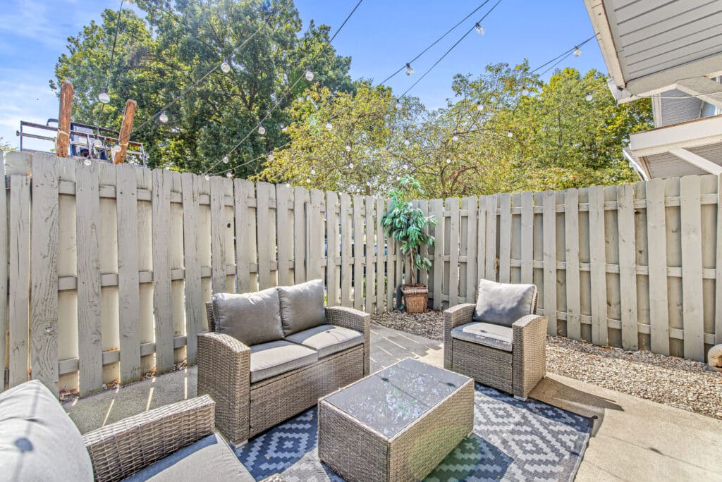 Outdoor patio area with wicker furniture, a patterned rug, potted plant, string lights overhead, and a wooden privacy fence.