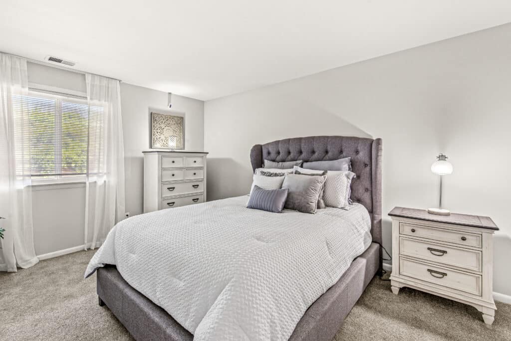 A neatly made bed with gray tufted headboard, two nightstands, a dresser, and a window with sheer curtains in a bright, neutral-toned bedroom.