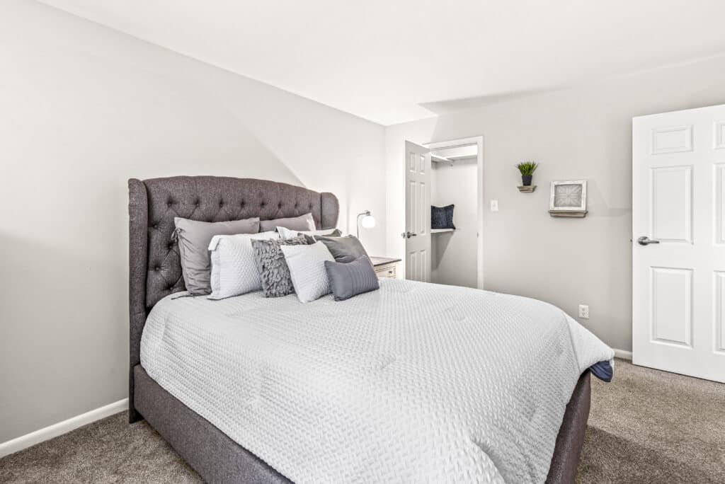 A neatly made bed with gray and white bedding is in a bright, minimalist bedroom with light walls, carpeted floor, and open doors leading to adjoining rooms.