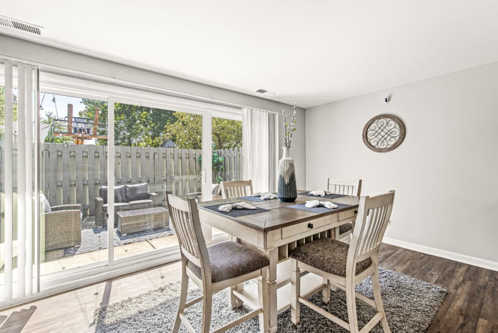 Bright dining area with a wooden table and four chairs, placed on a rug near sliding glass doors that open to a fenced outdoor patio with seating.
