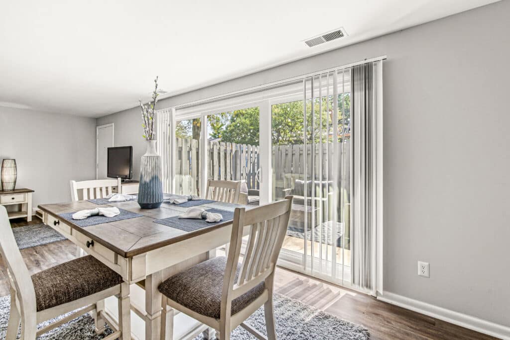 A dining area with a wooden table and chairs is next to large glass sliding doors that open to a fenced outdoor patio. Sunlight brightens the room through the doors.