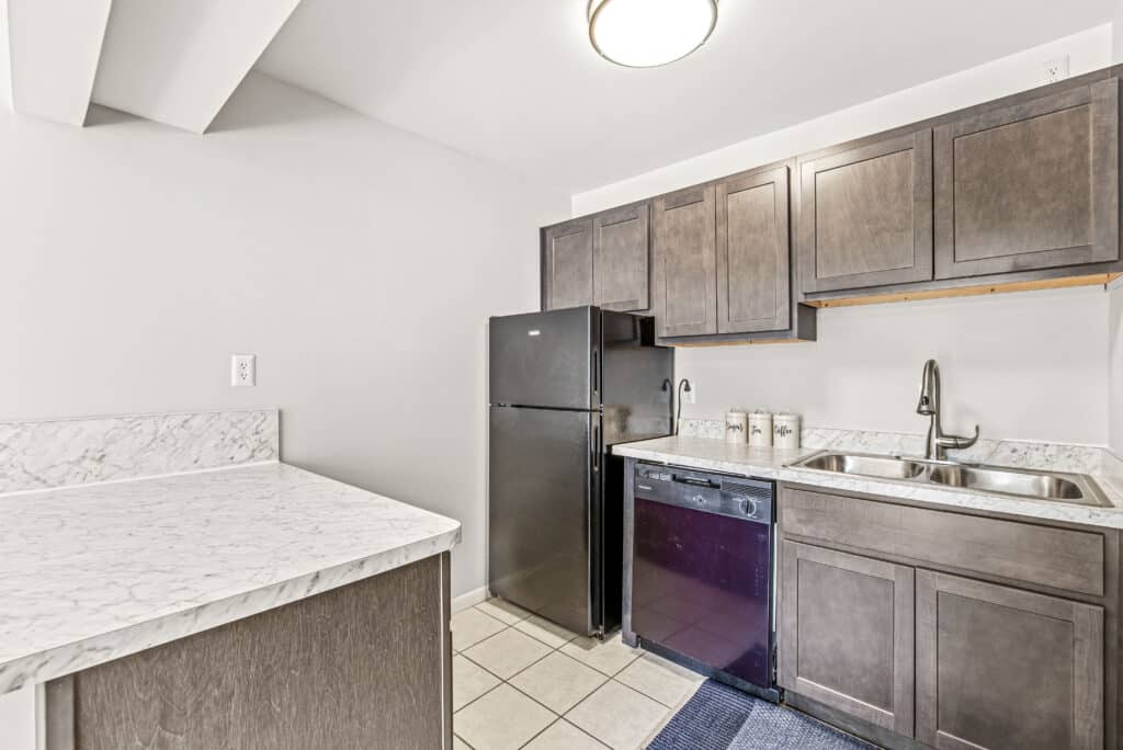 Modern kitchen with dark wood cabinets, black appliances, double sink, marble countertops, tile flooring, and a small blue rug.