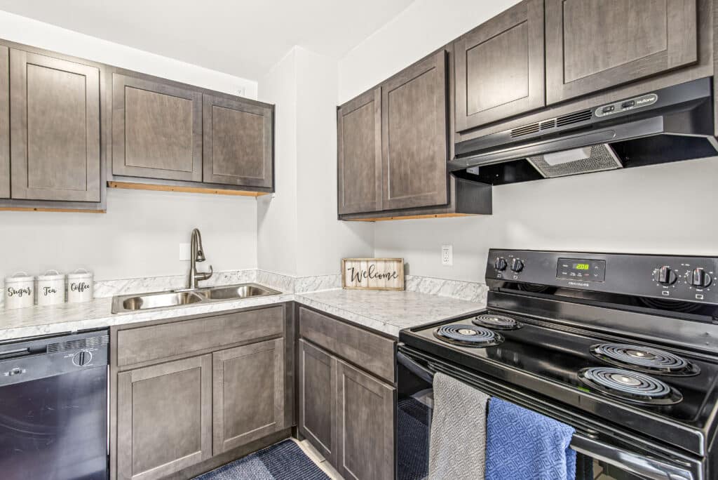 Modern kitchen with dark wood cabinets, electric stove, sink, dishwasher, and labeled jars for sugar, flour, and coffee on the countertop.