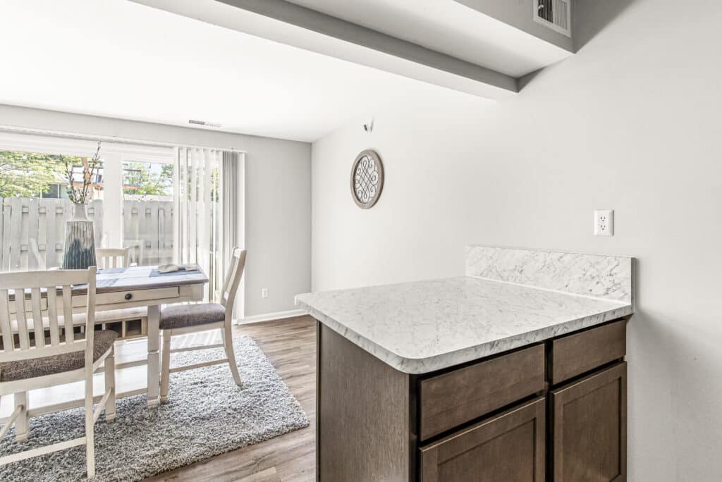 A modern dining area with a wooden table and chairs sits near a sliding glass door; a marble countertop and dark cabinets are in the foreground.