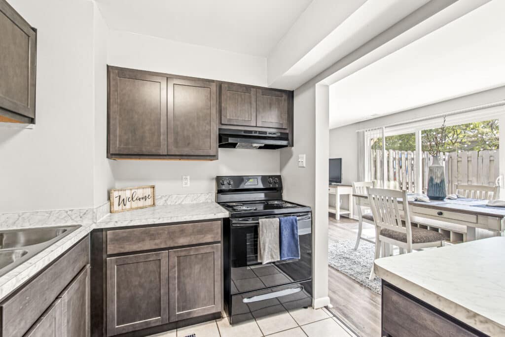 Modern kitchen with dark wood cabinets, electric stove, and marble countertops; adjoining dining area visible through open doorway with natural light.