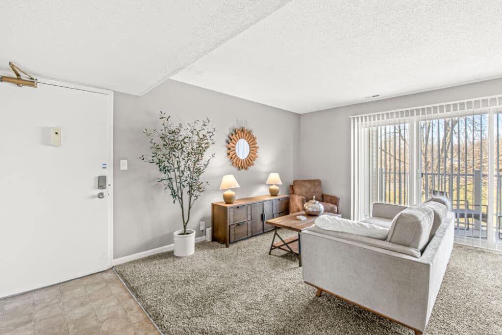 Bright living room with a gray sofa, brown armchair, wooden coffee table, sideboard with lamps, potted plant, and large window with vertical blinds showing trees outside.