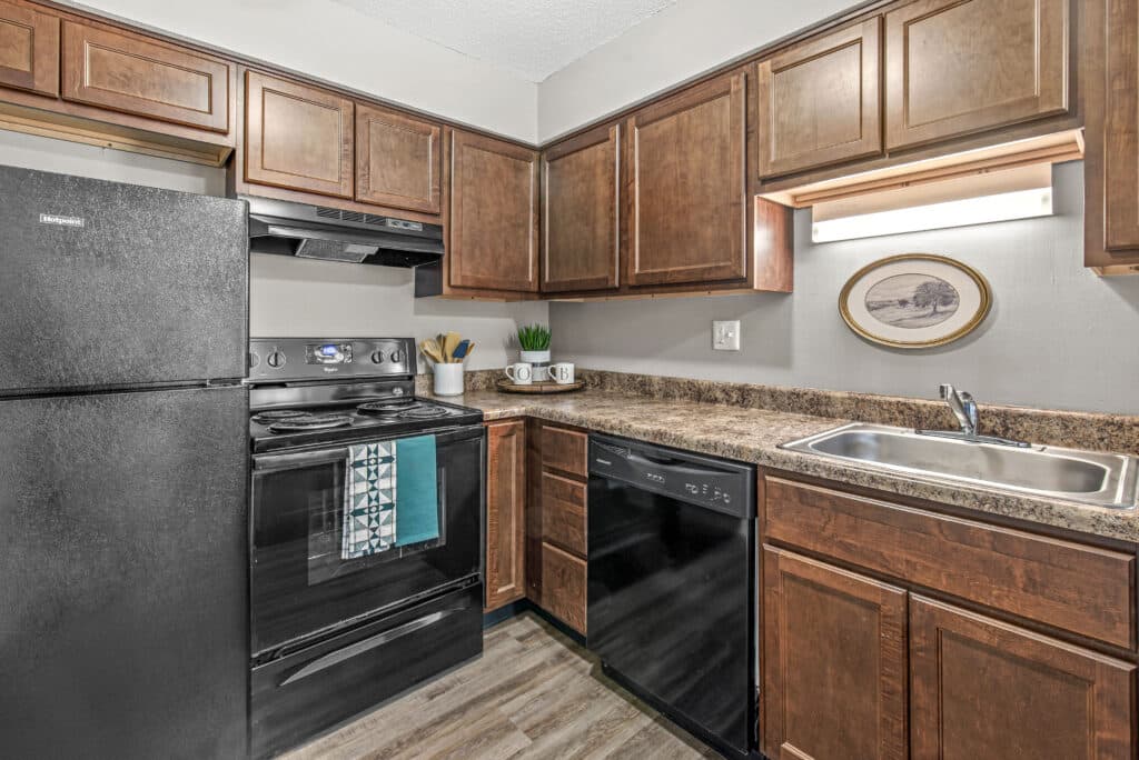 Compact kitchen with dark wood cabinets, black appliances, a double sink, and brown marbled countertops; small plant and dishes on the counter, framed art above the sink.