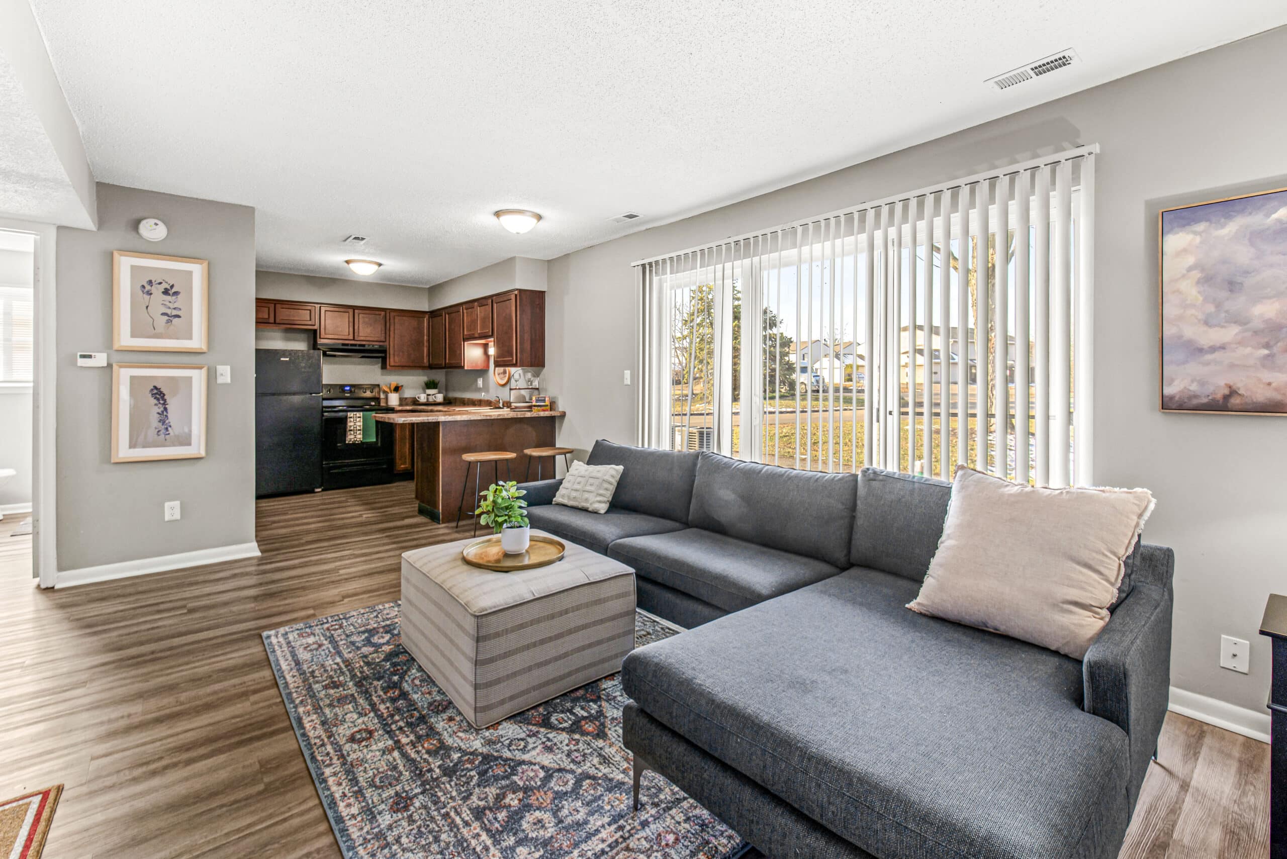 A modern living room with a gray sectional sofa, a small ottoman, a patterned rug, and a kitchen area with dark cabinets and black appliances in the background. Vertical blinds cover large windows.