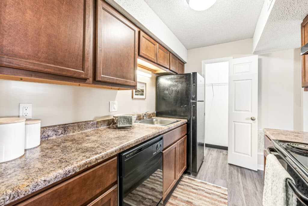 Galley kitchen with dark wood cabinets, black appliances, a double sink, marble-patterned countertop, and an open pantry door in the background.