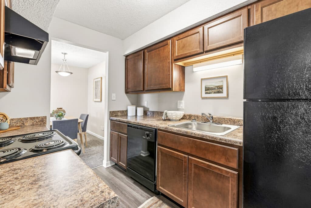 A galley kitchen with brown cabinets, black appliances, double sink, and marble-patterned countertops. Dining area with table and chairs visible through doorway.