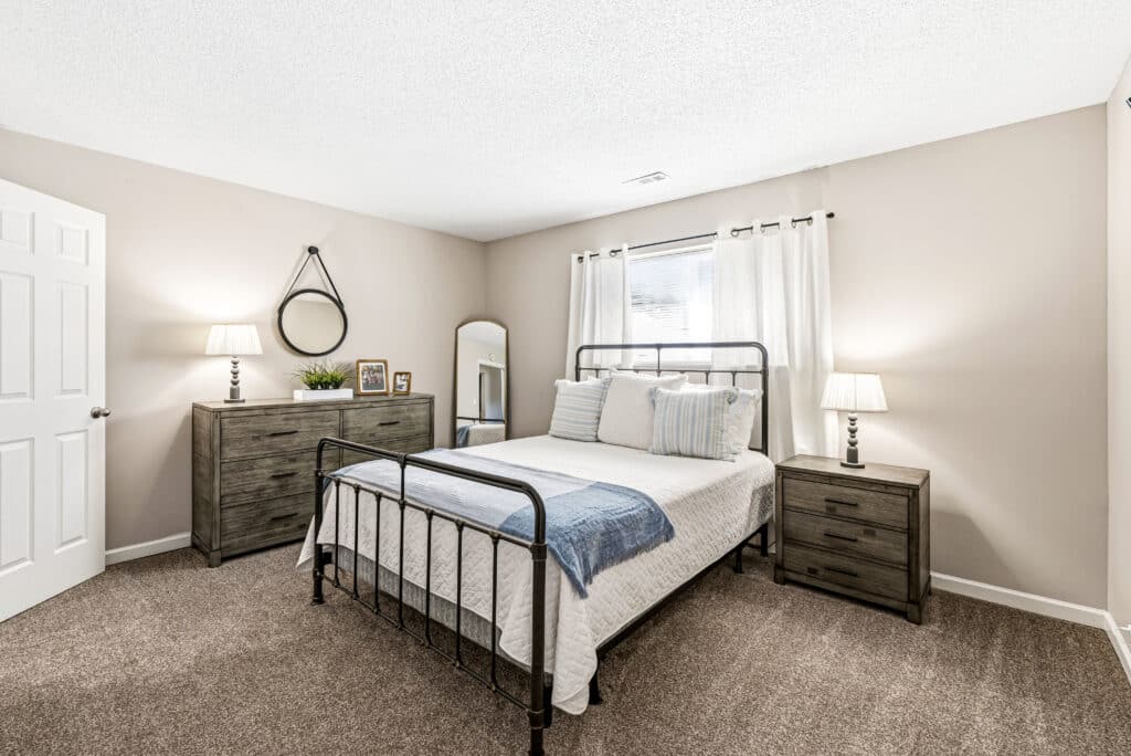 A bedroom with a metal bed frame, white bedding, blue accents, two nightstands with lamps, a dresser with decor, a circular mirror, a tall mirror, and a window with white curtains.