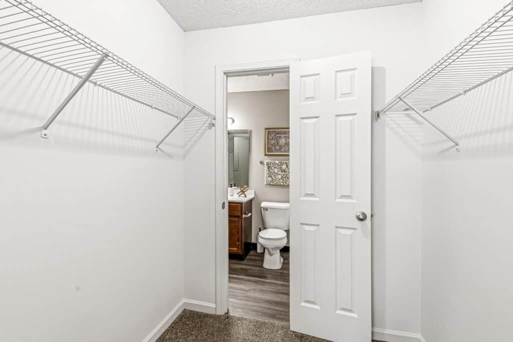 A walk-in closet with wire shelves on both sides, beige carpet flooring, and an open door leading to a bathroom with a toilet and vanity.