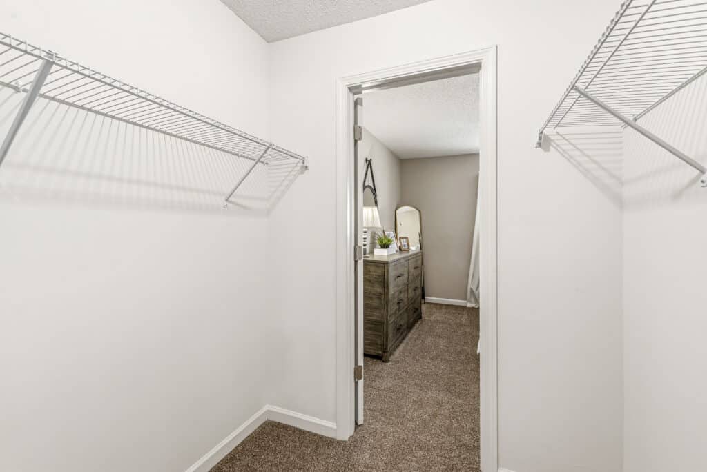 Empty walk-in closet with white walls and wire shelves, looking through an open door into a bedroom with a dresser, lamp, and mirror.