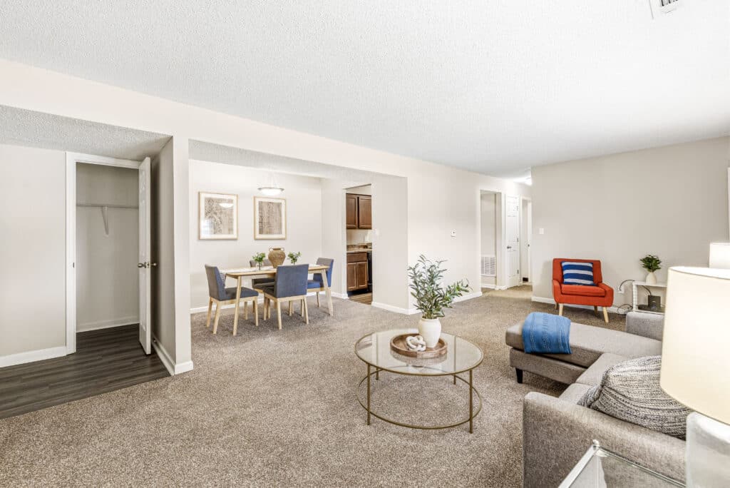 A spacious living and dining area with beige carpet, gray sofa, round glass coffee table, dining table with chairs, and an open doorway to the kitchen.