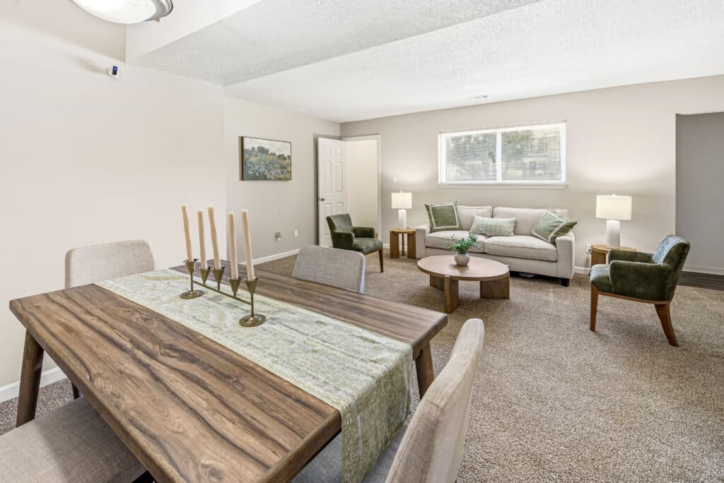 A living and dining area with a wooden table, upholstered chairs, a sofa, armchair, coffee table, lamps, and wall art in a carpeted room with neutral tones.