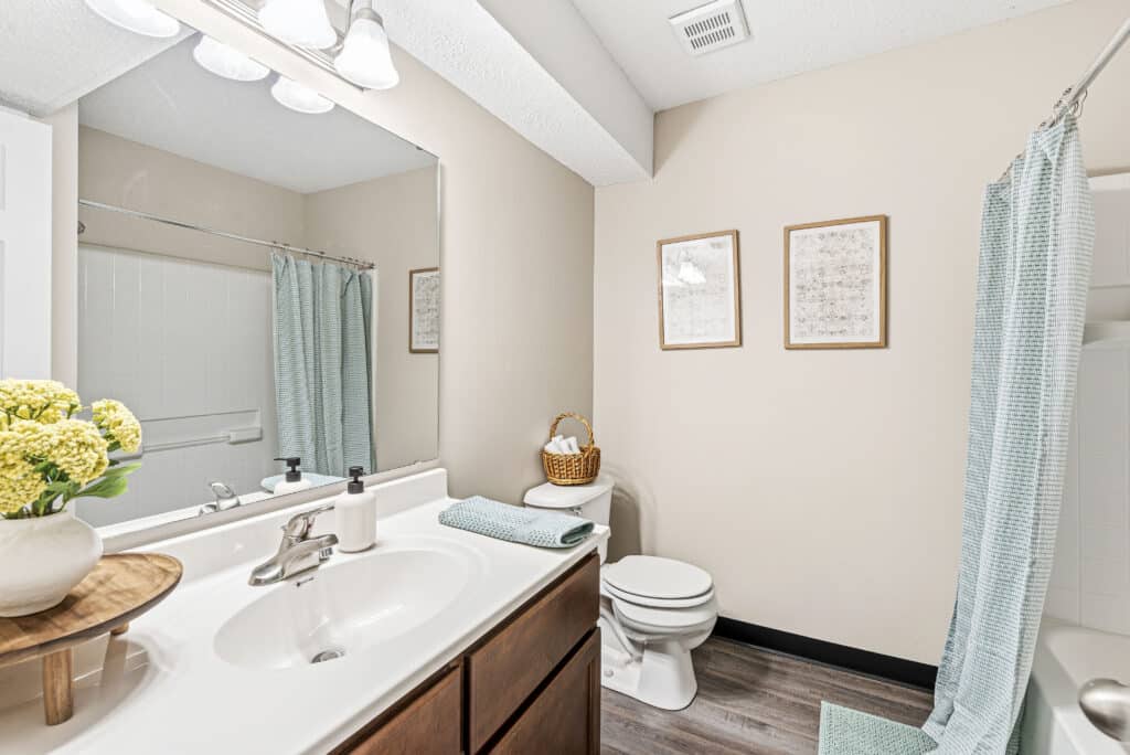 A clean bathroom with a sink and vanity, toilet, shower with a blue curtain, two framed pictures on the wall, and a vase of yellow flowers on the counter.