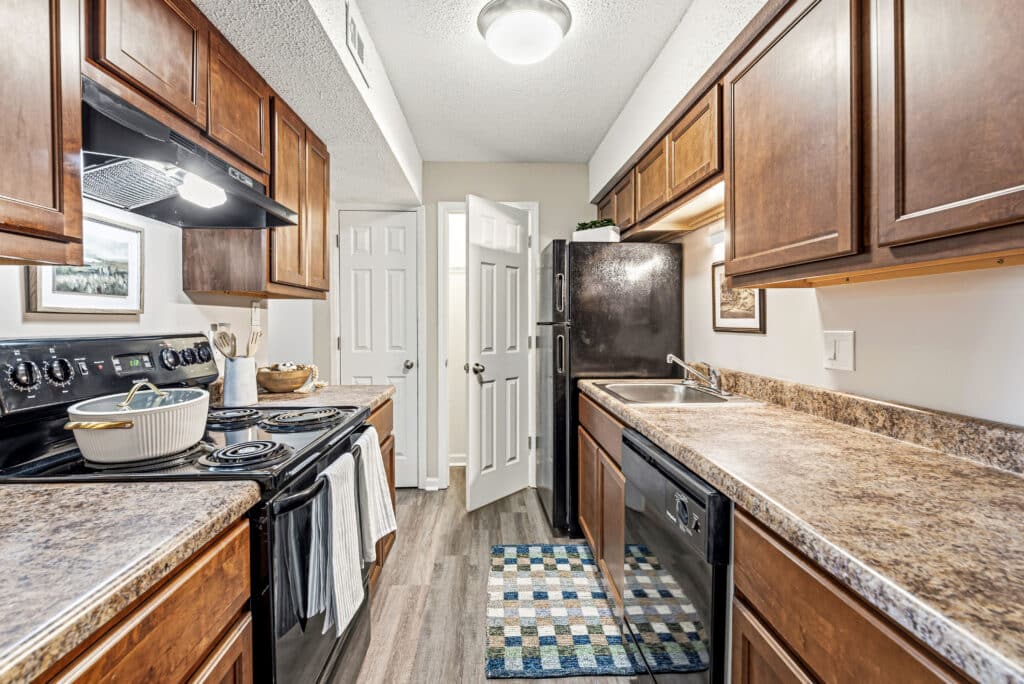 Narrow kitchen with wooden cabinets, black appliances, and a double sink. A patterned rug lies on the floor and a door is open at the far end of the room.