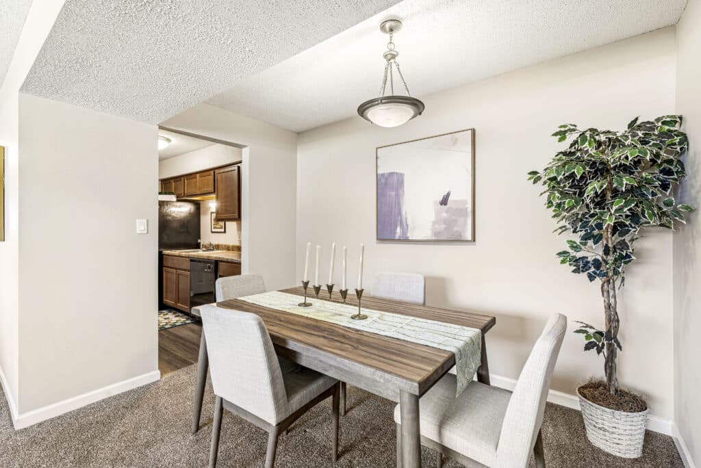 A small dining area with a wooden table, four upholstered chairs, a modern chandelier, a potted plant, and a wall painting, adjacent to a kitchen entrance.